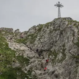 Grupa turystów wspina się po kamienistym zboczu w kierunku krzyża na Giewoncie. Trudno określić, ile dokładnie się idzie, ale widok jest imponujący.