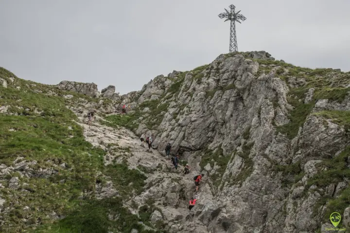 Grupa turystów wspina się po kamienistym zboczu w kierunku krzyża na Giewoncie. Trudno określić, ile dokładnie się idzie, ale widok jest imponujący.
